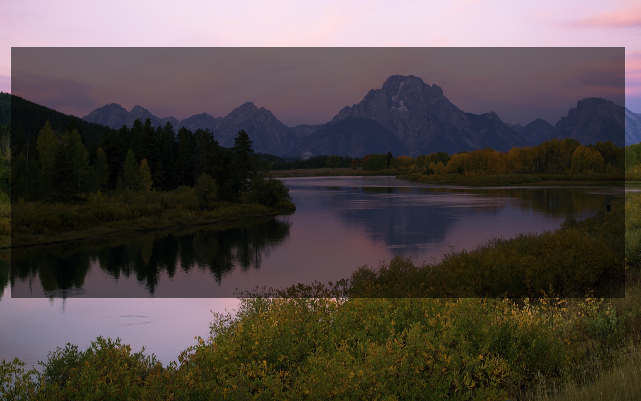 A photo of Mt. Moran, a large mountain in Grand Teton National Park, with the mountain reflected in the waters of Oxbow Bend. It is just after the sun has set behind the Teton Range and the sky is pink.
